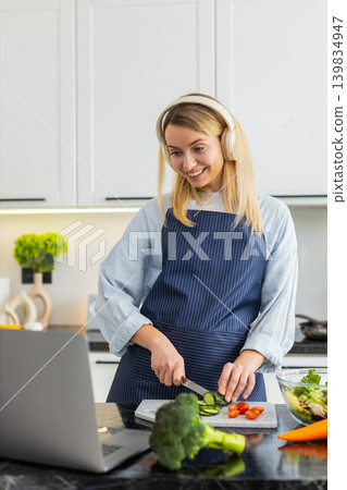 Young woman cutting cucumber for keto salad while watching recipe on laptop and wearing headphones Young woman cutting cucumber for keto salad while watching recipe on laptop and wearing headphones 139834947