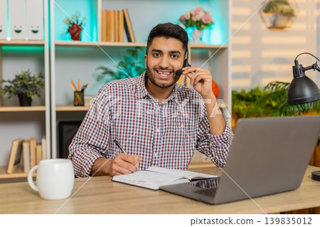 Indian man on home table working as support operator with headset writing notes during online call Indian man on home table working as support operator with headset writing notes during online call 139835012