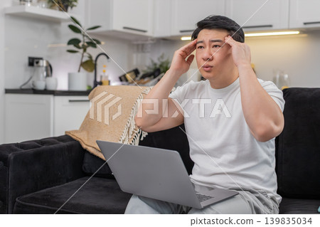 Asian man with laptop feels headache rubs temples closes eyes breathes slowly takes short work break Asian man with laptop feels headache rubs temples closes eyes breathes slowly takes short work break 139835034