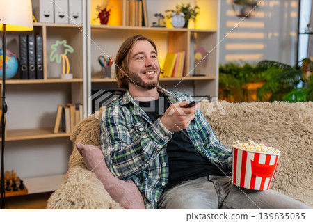 Young man on home sofa watching television with popcorn and remote smiling during fun evening rest Young man on home sofa watching television with popcorn and remote smiling during fun evening rest 139835035