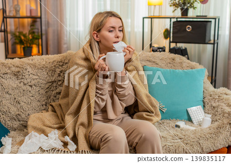 Woman covered with blanket on sofa, warming hands on tea cup, showing cold symptoms with tired look 139835117