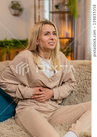 Adult woman holding stomach in pain on sofa at home, showing discomfort from food issues or cramps Adult woman holding stomach in pain on sofa at home, showing discomfort from food issues or cramps 139835358