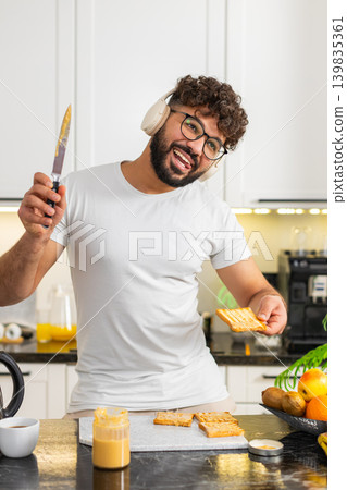 Arabian adult man dancing with headphones while making peanut butter toast breakfast in home kitchen Arabian adult man dancing with headphones while making peanut butter toast breakfast in home kitchen 139835361