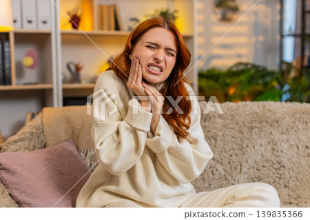 Young woman on home sofa touching cheek with tooth pain and showing sudden dental discomfort at home Young woman on home sofa touching cheek with tooth pain and showing sudden dental discomfort at home 139835366