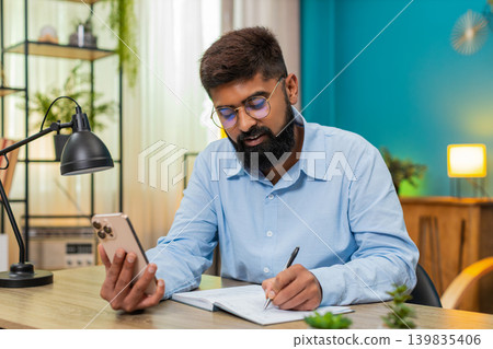 Indian man follows online lecture on smartphone nods writing notes focused distance learning class Indian man follows online lecture on smartphone nods writing notes focused distance learning class 139835406