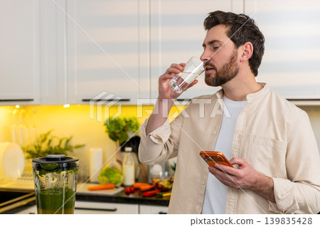 Middle-aged man drinks water checking smartphone in kitchen for weight loss diet hydration concept Middle-aged man drinks water checking smartphone in kitchen for weight loss diet hydration concept 139835428