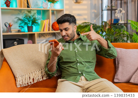 Indian man on home sofa arguing on smartphone speaker call with stop gesture and nervous reaction Indian man on home sofa arguing on smartphone speaker call with stop gesture and nervous reaction 139835433