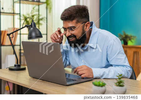 Indian man smiles after email reply on laptop nods celebrating project finish success indoors table Indian man smiles after email reply on laptop nods celebrating project finish success indoors table 139835468