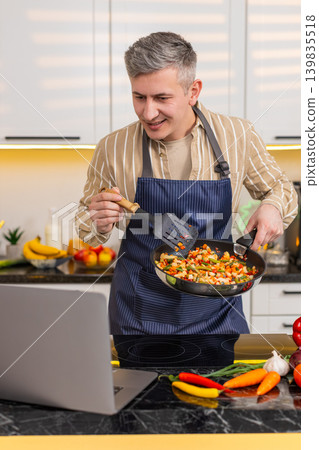 Middle-aged man frying vegetables while following laptop recipe for diet meal preparation in kitchen Middle-aged man frying vegetables while following laptop recipe for diet meal preparation in kitchen 139835518