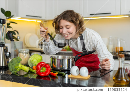 Young woman tasting soup in kitchen at home while cooking healthy meal with gentle smile and aroma 139835524