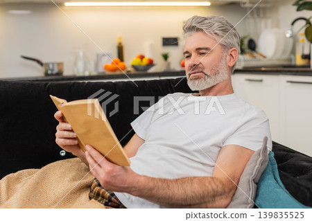 Middle-aged man enjoys reading book wrapped in plaid smiles and relaxes during quiet leisure time Middle-aged man enjoys reading book wrapped in plaid smiles and relaxes during quiet leisure time 139835525
