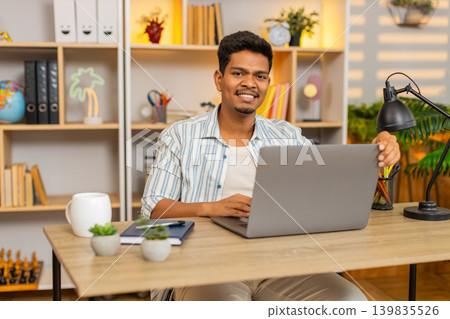 Indian man on home table opening laptop with cheerful smile and starting morning remote work indoors 139835526