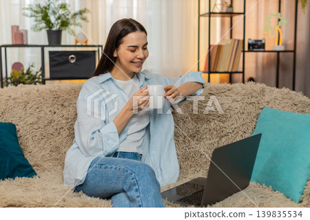 Young woman sitting on sofa happily watching online movie on laptop drinking tea relaxed enjoyment Young woman sitting on sofa happily watching online movie on laptop drinking tea relaxed enjoyment 139835534