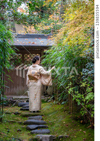 A woman in a kimono, seen from behind, walking through the gate of a garden surrounded by greenery. Kyoto Prefecture, Japan. 139835594