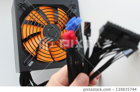 Computer Supply Block With High Performance Fan Behind Blurred Fingers Of Technician's Fingers Gripping A Wiring Harness 139835744