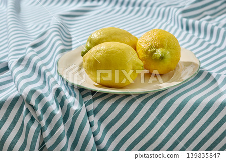 Fresh Lemons on a White Ceramic Plate on a Wrinkled Blue Striped Tablecloth. Front View Fresh Lemons on a White Ceramic Plate on a Wrinkled Blue Striped Tablecloth. Front View 139835847