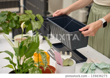 Woman prepares planter beside potted plants, gloves and garden tools on outdoor table Woman prepares planter beside potted plants, gloves and garden tools on outdoor table 139836273