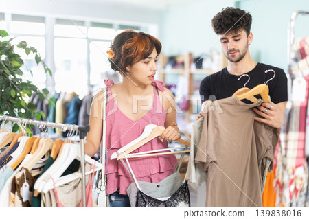 Girl spend time in shop and chooses tank top or dress, accompanying boyfriend choose t-shirt. 139838016