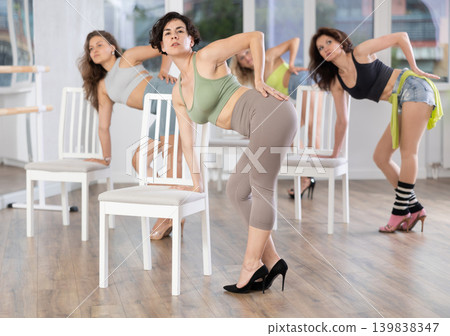 Energetic young female dancer practicing erotic movements of strip plastic near chair with other women in dance room Energetic young female dancer practicing erotic movements of strip plastic near chair with other women in dance room 139838347