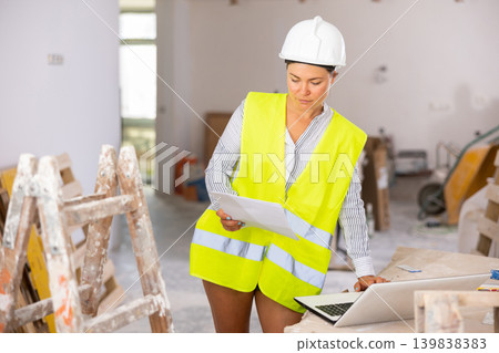 Female foreman in a protective helmet and a yellow vest checks the execution of repair work using laptop Female foreman in a protective helmet and a yellow vest checks the execution of repair work using laptop 139838383