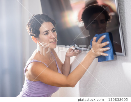 Positive young brunette woman cleaning dust from TV screen Positive young brunette woman cleaning dust from TV screen 139838605