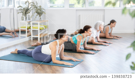 Group of women practicing pigeon pose of yoga on mat in fitness room Group of women practicing pigeon pose of yoga on mat in fitness room 139838723