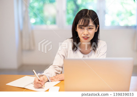 一位年輕的日本女性一邊在電腦前工作，一邊在筆記本上做筆記。 139839033
