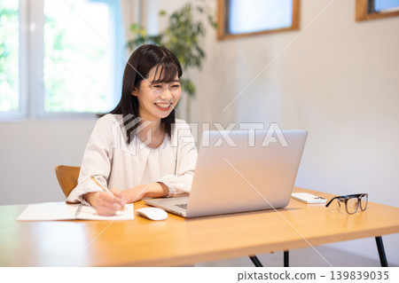 一位年輕的日本女性一邊在電腦前工作，一邊在筆記本上做筆記。 139839035