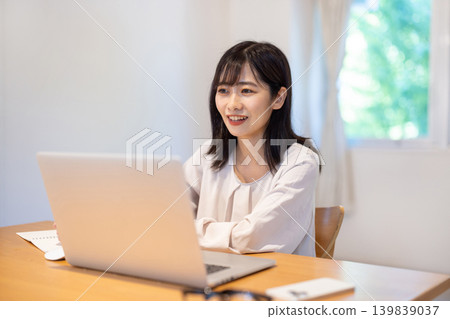 A young Japanese woman taking notes in a notebook while working on her computer. A young Japanese woman taking notes in a notebook while working on her computer. 139839037