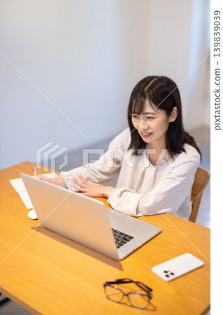 一位年輕的日本女性一邊在電腦前工作，一邊在筆記本上做筆記。 139839039