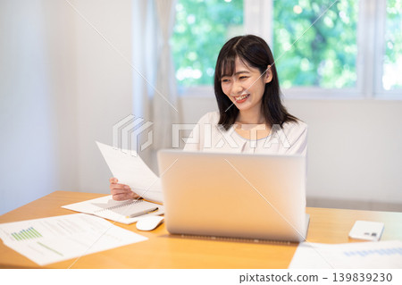 A young Japanese woman checking documents while working on her computer. A young Japanese woman checking documents while working on her computer. 139839230