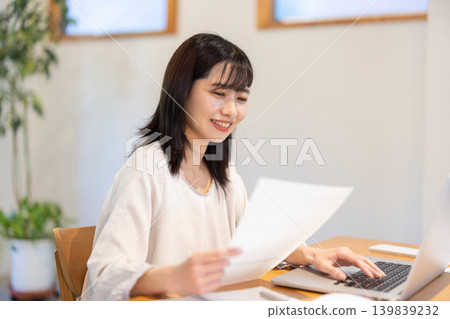 A young Japanese woman checking documents while working on her computer. A young Japanese woman checking documents while working on her computer. 139839232