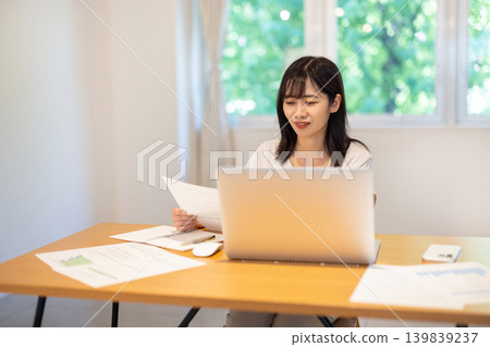 A young Japanese woman checking documents while working on her computer. A young Japanese woman checking documents while working on her computer. 139839237