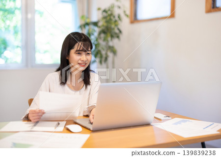 A young Japanese woman checking documents while working on her computer. A young Japanese woman checking documents while working on her computer. 139839238