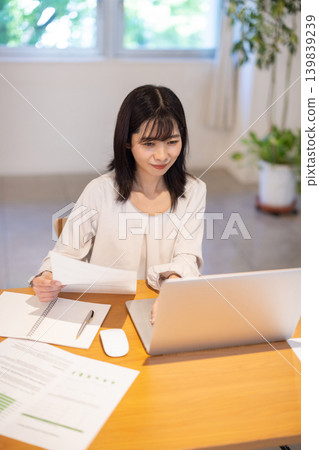 A young Japanese woman checking documents while working on her computer. A young Japanese woman checking documents while working on her computer. 139839239