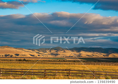 Expansive golden grasslands meet rolling hills under a dynamic sky. A long wooden fence crosses the foreground Expansive golden grasslands meet rolling hills under a dynamic sky. A long wooden fence crosses the foreground 139839490