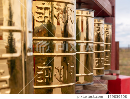 Golden Buddhist prayer wheels stand on a rustic wooden shelf outdoors. Each wheel has detailed engravings and reflects the overcast daytime sky Golden Buddhist prayer wheels stand on a rustic wooden shelf outdoors. Each wheel has detailed engravings and reflects the overcast daytime sky 139839508