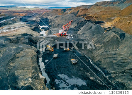 Massive dragline excavator at work in a sprawling open-pit coal mine. Several haul trucks transport excavated material Massive dragline excavator at work in a sprawling open-pit coal mine. Several haul trucks transport excavated material 139839515
