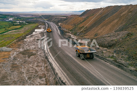 Two large yellow dump trucks drive on a wide dirt road within an open pit mine 139839525