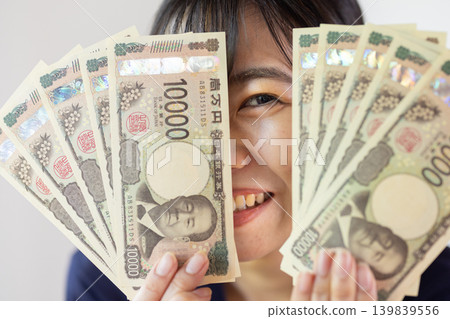 A young Japanese woman smiles, holding ten 10,000 yen bills spread out. A young Japanese woman smiles, holding ten 10,000 yen bills spread out. 139839556