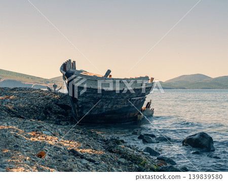 A decaying wooden boat, broken and weathered, lies abandoned on a stone shore A decaying wooden boat, broken and weathered, lies abandoned on a stone shore 139839580