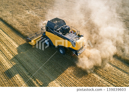 Aerial view captures a large yellow combine harvester actively cuts ripe golden grain in a vast agricultural field on a clear sunny summer day 139839603