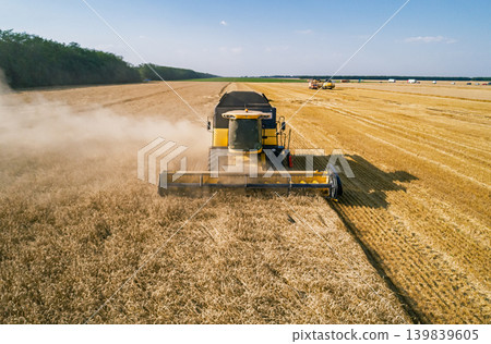 Aerial view captures a large yellow combine harvester actively cuts ripe golden grain in a vast agricultural field on a clear sunny summer day Aerial view captures a large yellow combine harvester actively cuts ripe golden grain in a vast agricultural field on a clear sunny summer day 139839605