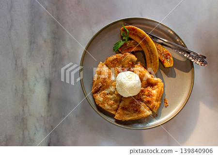 Banana roti topping with vanilla ice cream is arranged on a modern style plate on the table. Top view. Banana roti topping with vanilla ice cream is arranged on a modern style plate on the table. Top view. 139840906