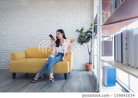 Young woman smartphone frustration sofa, casual outfit indoors home office, modern living room bookshelf background plant 139841034