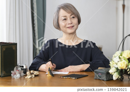 A senior woman paints picture postcards in her living room. 139842021