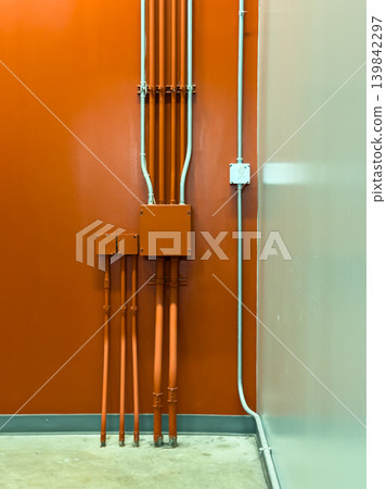 Pipes and electrical setup against a bright orange wall in a building hallway during daylight hours Pipes and electrical setup against a bright orange wall in a building hallway during daylight hours 139842297