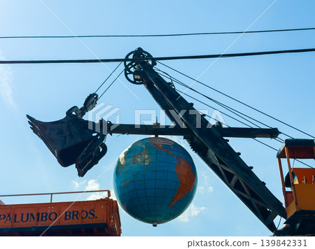 Workers use a crane to lift a large globe in a construction area under a clear blue sky during the day 139842331