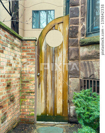 Wooden door stands at the entrance of a brick wall, leading to a small outdoor space in an urban neighborhood where buildings surround it Wooden door stands at the entrance of a brick wall, leading to a small outdoor space in an urban neighborhood where buildings surround it 139842338