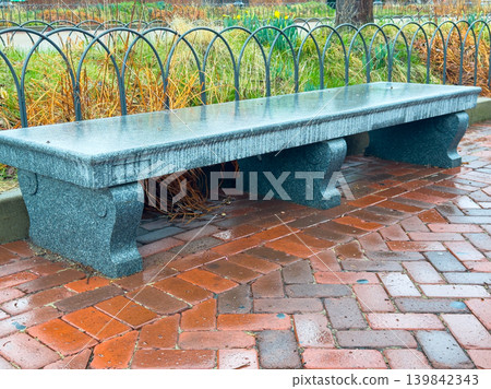 Sturdy stone bench placed on brick pathway in park surrounded by greenery on a cloudy day 139842343